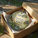 A decorative ornament in a brown box, featuring a scenic landscape with a lake, trees, and a bridge.