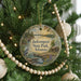 A round ornament hanging on a Christmas tree, featuring a scenic landscape with a river, cabin, and trees.
