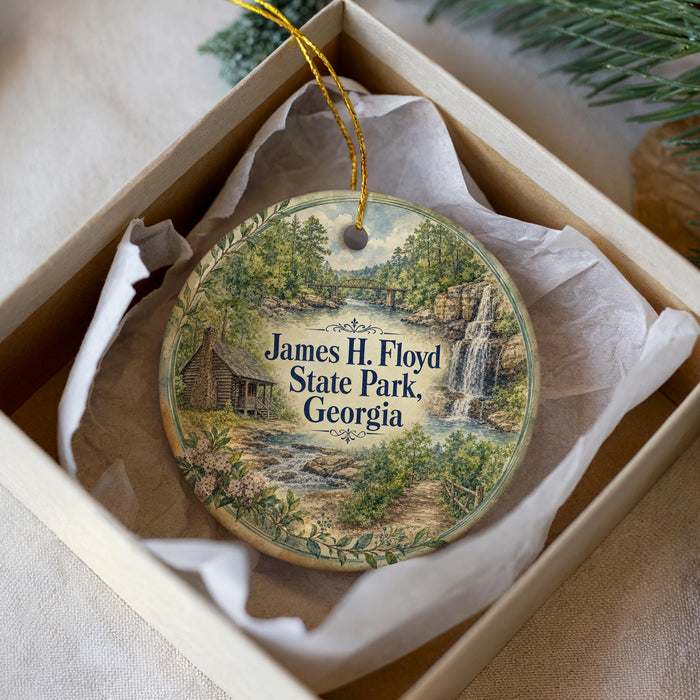 A decorative ornament in a box, featuring a scenic landscape with a waterfall and a cabin, with the text "James H. Floyd State Park, Georgia" printed on it.