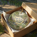 A decorative ornament in a box, featuring a scenic landscape with a waterfall and greenery, with the text "James H. Floyd State Park Georgia" printed on it.