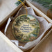 A round ornament with a painting of a historic plantation house and a pond, placed in a white box with a gold string.
