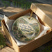 A decorative ornament in a box, featuring a scenic landscape with mountains, trees, and a river.