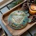 A decorative ornament featuring a tropical scene with a lighthouse, palm trees, and a coral reef, placed on a wooden tray alongside pine cones and a lit candle.