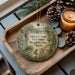 A wooden tray with a decorative ornament featuring a scenic trail and bridge, surrounded by pine cones and a lit candle.