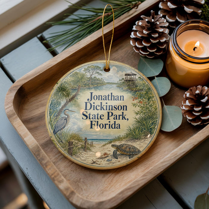 A decorative ornament featuring a bird and palm trees, with the text "Jonathan Dickinson State Park, Florida" displayed on it, placed on a wooden tray alongside pine cones and a lit candle.