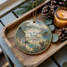 A wooden tray with a decorative ornament featuring a scenic landscape of Julia Pfeiffer Burns State Park, California, and a squirrel. The ornament is surrounded by pine cones and a lit candle, creating a cozy and festive atmosphere.