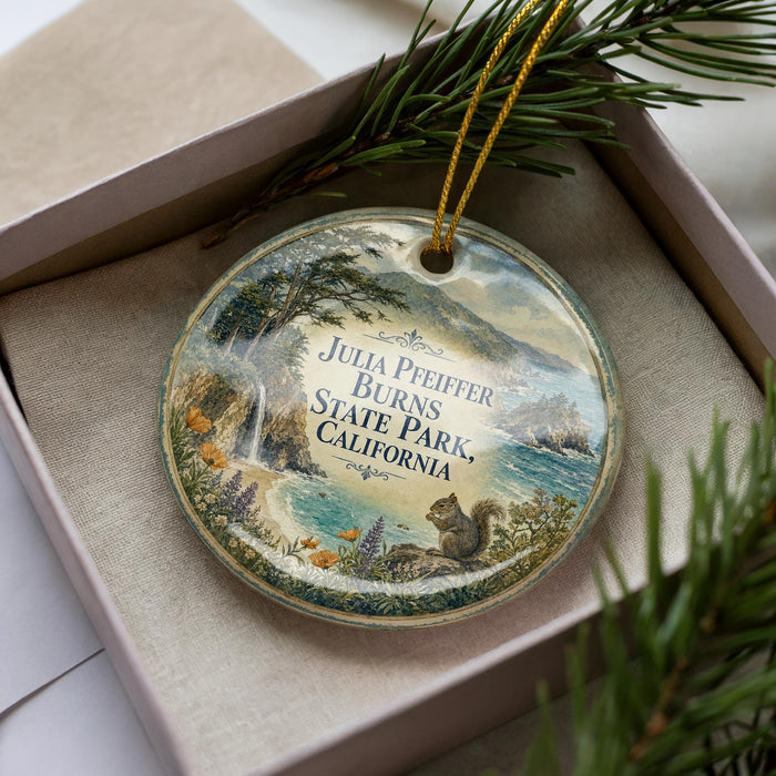 A decorative ornament in a box, featuring a scenic landscape with a squirrel, pine trees, and a waterfall.