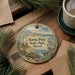 A decorative ornament with a lighthouse and a bird, featuring the text "Kaena Point State Park, Hawaii" on it, is placed on a wooden surface alongside a cup of coffee and some pine branches.