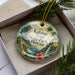 A decorative ornament with a tropical landscape design, featuring palm trees, a waterfall, and a hibiscus flower, is displayed in a box with pine branches.