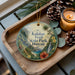 A circular ornament with a tropical landscape design, featuring palm trees, a waterfall, and flowers, is displayed on a wooden tray alongside pine cones and a lit candle.