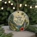 A circular ornament with a tropical landscape design, featuring palm trees, a waterfall, and a hibiscus flower, hangs in front of a blurred background with Christmas lights.