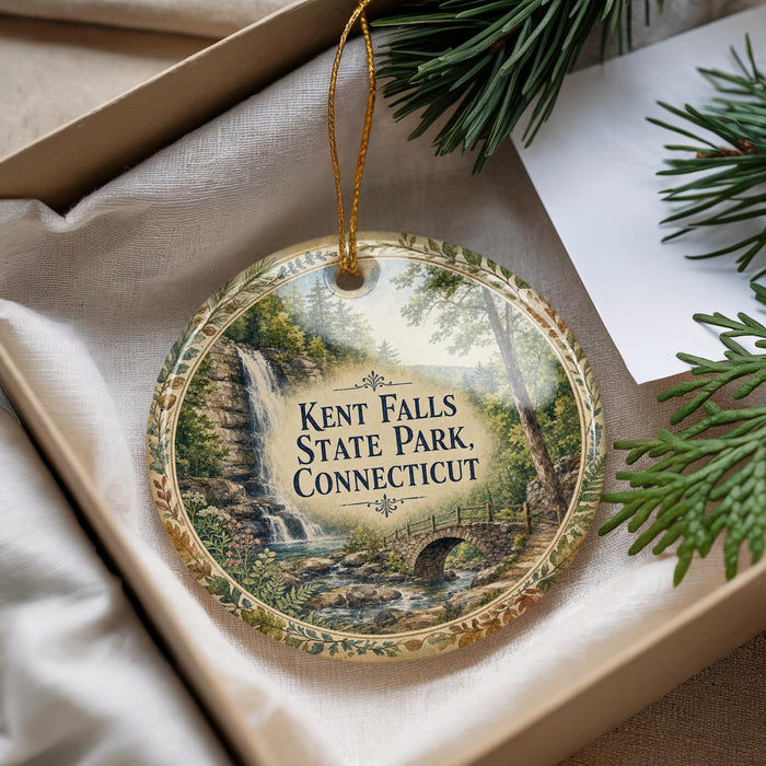 A decorative ornament with a waterfall and bridge image, hanging from a string, placed inside a box with pine branches.