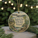 A round ornament with a scenic illustration of Kentucky, featuring a bridge, houses, and a lake, is displayed against a blurred background of Christmas lights.