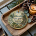 A decorative ornament featuring a scenic image of Kilen Woods State Park in Minnesota, with pine cones and a lit candle on a wooden tray.