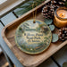 A decorative ornament with a painting of a pond and surrounding scenery, placed on a wooden tray alongside pine cones and a lit candle.