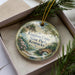 A decorative ornament in a box, featuring a scenic landscape with a bridge and trees, with the text "Kingdom Come State Park, Kentucky" printed on it.