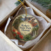 A decorative ornament in a box, featuring a scenic landscape with mountains, trees, and a waterfall.