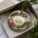 A decorative ornament with a landscape scene featuring mountains, pine trees, and a waterfall, placed inside a box with a pine branch.