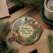 A circular ornament with a scenic image of a park, featuring a mountain landscape, pine trees, and red flowers, hangs on a wooden surface.