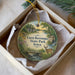 A round ornament with a scenic landscape featuring a river, a bridge, and a deer, placed in a box with a white tissue paper lining.