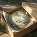 A round ornament in a box, featuring a scenic landscape with a river, trees, and a deer.