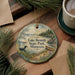 A decorative ornament featuring a lake, pine trees, and a bird, with the text "Lake Bemidji State Park, Minnesota" printed on it. The ornament is placed on a wooden surface, surrounded by pine branches and a cup of coffee.
