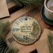 A decorative ornament featuring a lake, pine trees, and a dock, with the text "Lake Carlos State Park, Minnesota" displayed on it. The ornament is placed on a wooden surface, surrounded by pine branches and a cup of coffee.