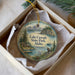 A decorative ornament in a box, featuring a scenic landscape with a lake, mountains, and a cabin, along with a bird perched on a rock.