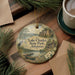 A decorative ornament featuring a scenic landscape of Lake Charles State Park in Arkansas, with a boat on the water and a cabin in the background. The ornament is hanging from a string and is placed on a wooden surface, surrounded by pine branches and a cup of coffee.