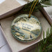 A decorative ornament with a lake, birds, and greenery, placed in a box with pine branches.