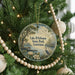 A round ornament hanging on a Christmas tree, featuring a scenic landscape with a lake, trees, and birds.