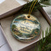 A decorative ornament with a scenic landscape of Lake Dardanelle State Park in Arkansas, featuring a deer and a house, is displayed in a box with pine branches.