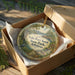 A decorative ornament in a wooden box, featuring a scenic landscape with a lake, mountains, and greenery.