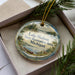A decorative ornament with a lake and forest scene, featuring a canoe and a deer, is displayed in a box with pine branches.