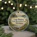 A round ornament with a scenic lake and forest landscape, featuring a boat and a deer, is displayed in front of a blurred background with Christmas lights.
