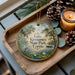 A decorative ornament with a serene scene of Lake Kissimmee State Park, Florida, featuring palm trees, a bird, and a pond. The ornament is placed on a wooden tray, with pine cones and a lit candle nearby.