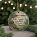 A round ornament with a scenic lake and surrounding landscape, featuring a bird and a canoe, is displayed in front of a blurred background with Christmas lights.