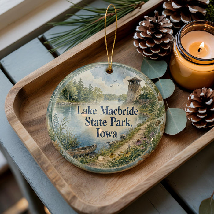 A decorative ornament featuring a scenic view of Lake Macbride State Park, Iowa, with a small boat on the water and a lighthouse in the distance. The ornament is displayed on a wooden tray, surrounded by pine cones and a lit candle.