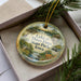 A decorative ornament with a lake scene and text, placed in a box with pine branches.