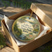 A decorative ornament in a box, featuring a scenic landscape with a lake, trees, and wildlife.