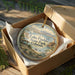 A decorative ornament in a box, featuring a scenic landscape with a lake, mountains, and a small boat, along with the text "Lake Pueblo State Park, Colorado".