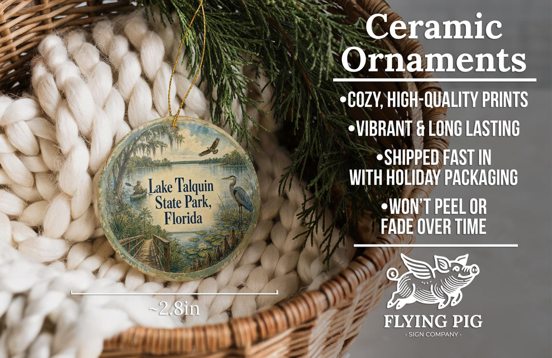 Ceramic ornaments, featuring a round ornament with a landscape scene and text, are displayed in a basket with pine branches.