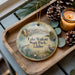 A wooden tray with a decorative ornament featuring a scenic lake and mountains, along with pine cones and a lit candle.