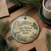 A decorative ornament with a scenic lake and mountains image, featuring a fish and a bird, is displayed on a wooden surface alongside a cup of coffee and some pine branches.