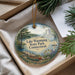 A decorative ornament with a scenic lake and surrounding landscape, featuring a boat and pine trees, is displayed in a box with pine branches.