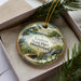 A decorative ornament with a scenic image of Louisiana State Park, featuring a bird, a bridge, and a house, is displayed in a box with pine branches.