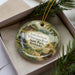 A decorative ornament in a box, featuring a scenic landscape with a cabin, trees, and a waterfall.