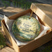 A round ornament in a brown box, featuring a scenic landscape with a lake, trees, and a bench.