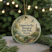 A round ornament with a lighthouse and a path, surrounded by greenery, is displayed in front of a blurred background with twinkling lights.