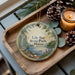 A wooden tray with a decorative ornament featuring a scenic landscape of a lake, pine trees, and mountains, along with pine cones and a lit candle.
